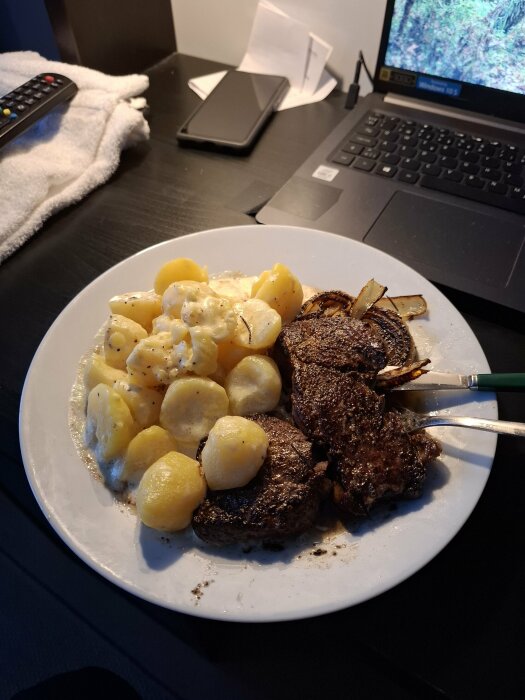 Tallrik med entrecote och potatis framför en bärbar dator, fjärrkontroll och mobiltelefon på ett skrivbord.