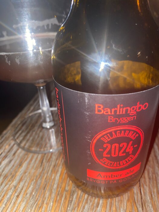 Flaska med Barlingbo Bryggeri Amber ale och ett glas öl på ett träbord. Etikett i rött med "Delägarnas Specialbrygd 2024".