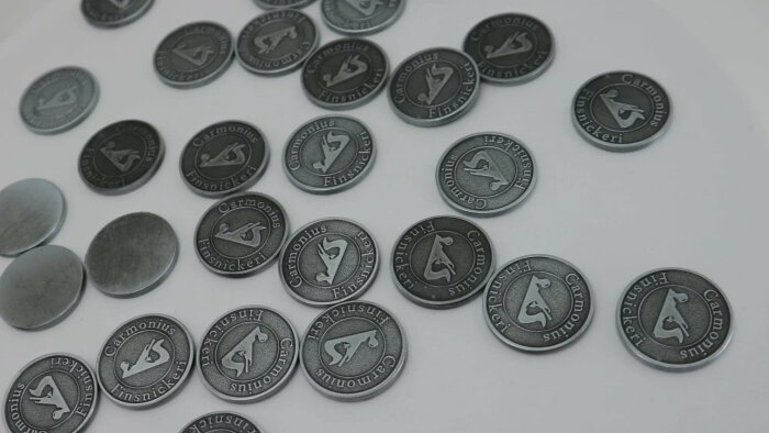 Små, runda metallbrickor med gravyr, cirka 20 mm i diameter. De har en logotyp och inskriptionen "Carmonius Finsnickeri".