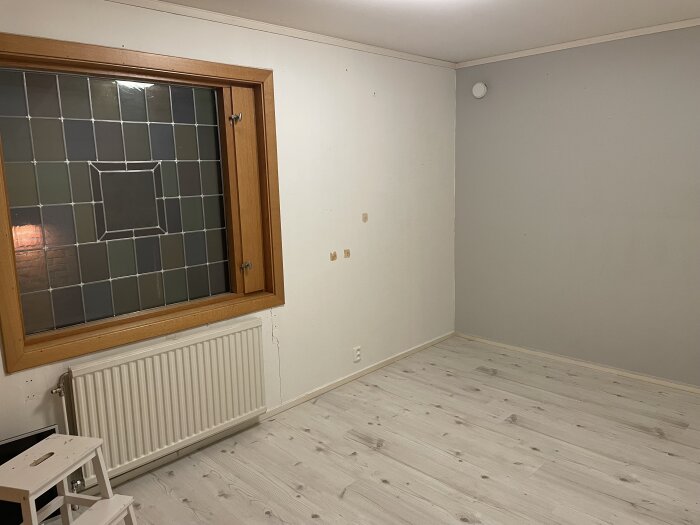 Tomt rum med ljus trägolv, ljusa väggar och ett stort fönster med träram och glasruta, sett innan renovering.