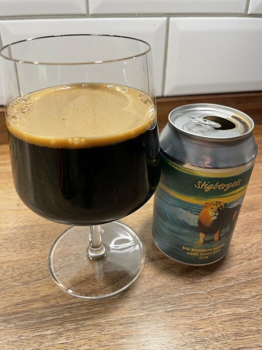 Ett glas stout bredvid en burk från Stigbergets bryggeri, märkt med "Big Bourbon Barrel Aged Stout 2021" på ett träbord.