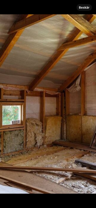 Ovanvåning under renovering med synliga takbjälkar, isoleringsmaterial på väggar och golv, samt ett fönster.