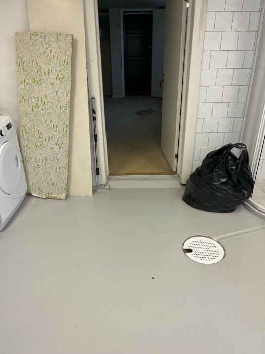 Källare under renovering med vitmålade väggar, golvbrunn, och borttagen heltäckningsmatta. Dörröppning till annan del av källaren syns.