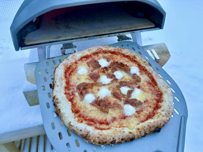 Hembakad pizza på en perforerad spade framför en ugn utomhus i snön.