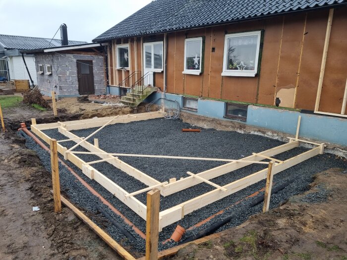 Pågående husrenovering med riven fasad och tilläggsisolering. Grunden för utbygge syns med träformar och grus på marken.