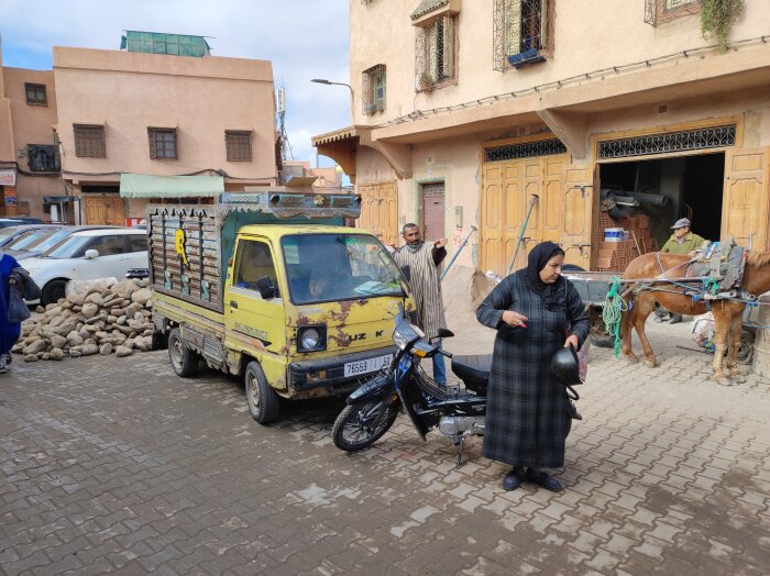 Gul lastbil, motorcykel och trava stenar i Marrakech, med människor och en häst i bakgrunden på ett gata.