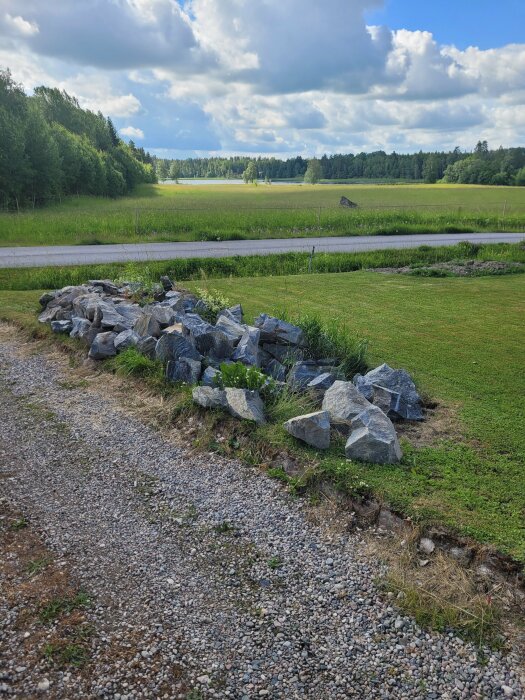 Hög med stenar vid grusväg och grön gräsmatta med skog och fält i bakgrunden under molnig himmel.