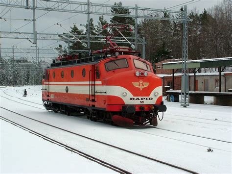 Rött och vitt elektriskt lokomotiv med texten "RAPID" under vintertid, stationerat på en järnväg med omgivande snötäckt landskap.