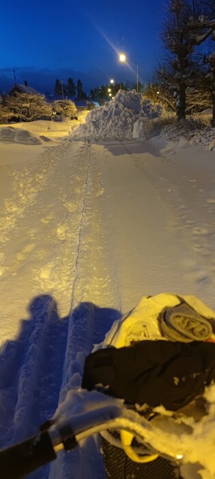 Snötäckt väg med stora snöhögar under kvällstid, sett från en cykels handtag med handskar, upplyst av gatubelysning.