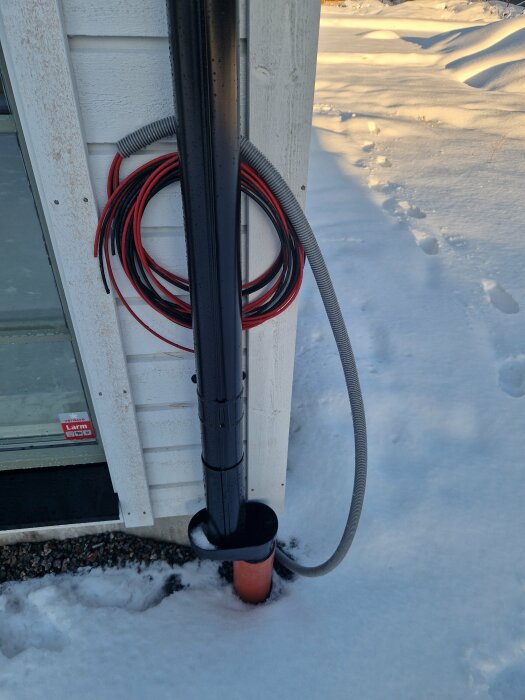 Svart och röd kabel, samt flexslang fästa på husvägg med snö på marken; installation för solceller till växelriktare.