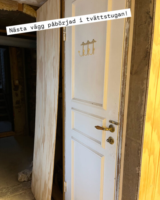 Vit dörr i tvättstuga med nymonterad obehandlad vägg av plywood till vänster, krok på dörren och text "Nästa vägg påbörjad i tvättstugan".