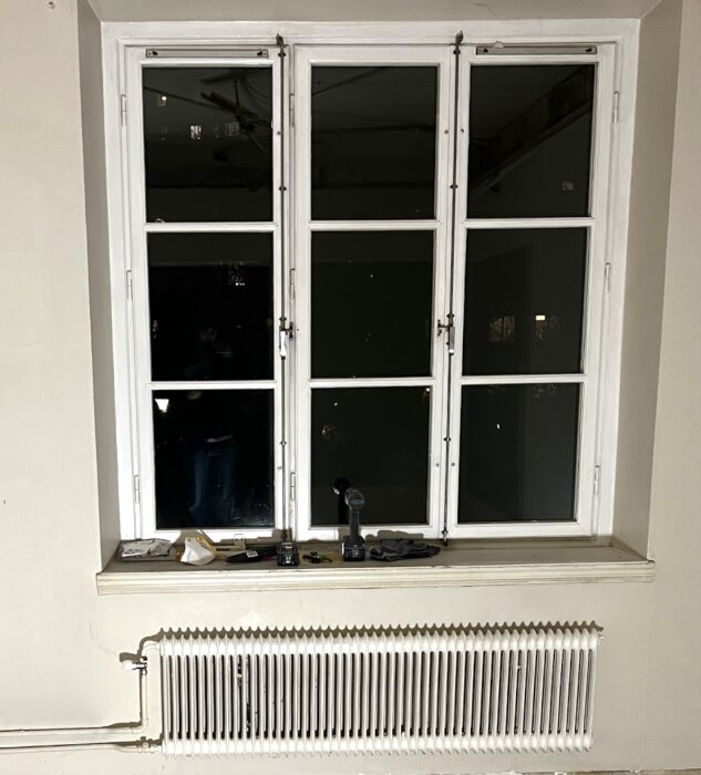 Stor fönsterinstallation med vita spröjs, mörk utsida. Verktyg och ventilationsrelaterade material på fönsterbrädan ovanför ett vitt element.