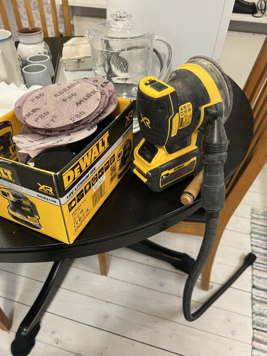 Sladdlös DeWalt slipmaskin med sandpapper på ett bord, bredvid vattenkokare och muggar.