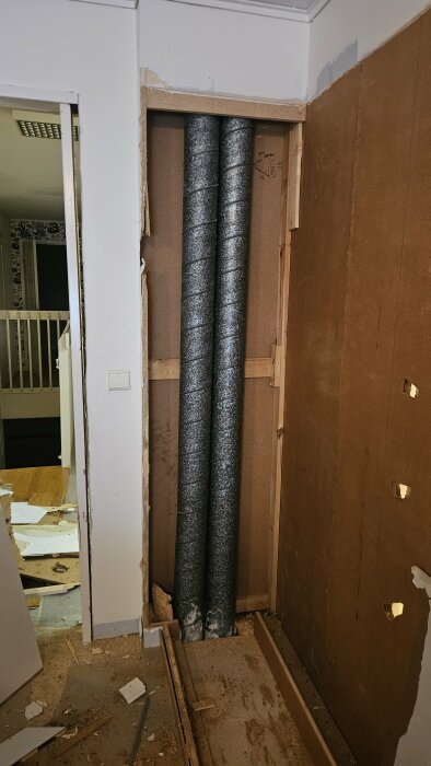Två synliga ventilationsrör i ett halvrivet väggutrymme under en renovering, med byggdamm och träbitar på golvet.