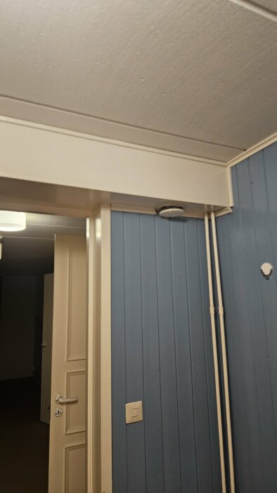 Bild av en dörröppning i ett rum med blå träpanelvägg och vit taklist, visar två vertikala ventrör nära taket.