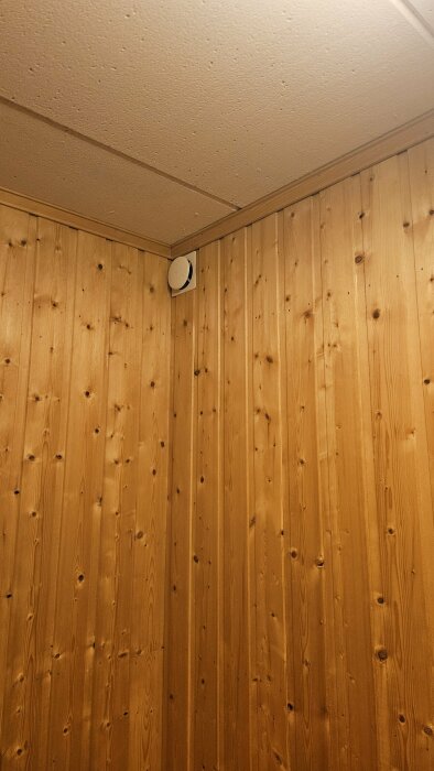 Vägg med träpanel i hörn av rum, vit rund ventilationsöppning nära taket.