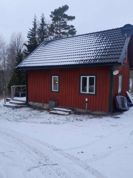 Röd stuga med svart tak i vintermiljö, omgiven av snö och skog. En parabolantenn är monterad på väggen.