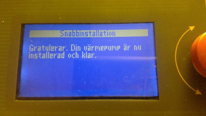 Display med meddelandet "Snabbinstallation: Gratulerar. Din värmepump är nu installerad och klar," mot blå bakgrund.