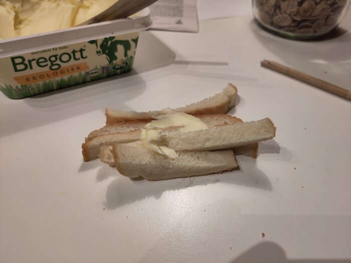 Skivat bröd med smör bredvid ett paket ekologiskt Bregott på ett bord, för kvällsfika och förberedelse till smörgåstårta.