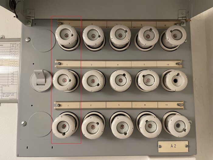 Bild på ett gammalt proppskåp med vita porslinssäkringar, vissa märkta med 10 A. Höljet är öppet med säkringar synliga i rader.