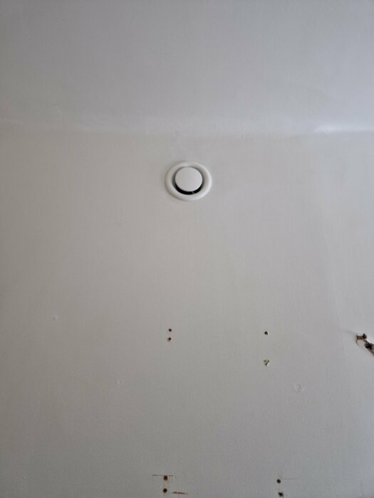 Ventilationsutlopp i tak med rund täckplatta, omgiven av vitmålad yta med några skruvhål och en liten spricka i närheten.