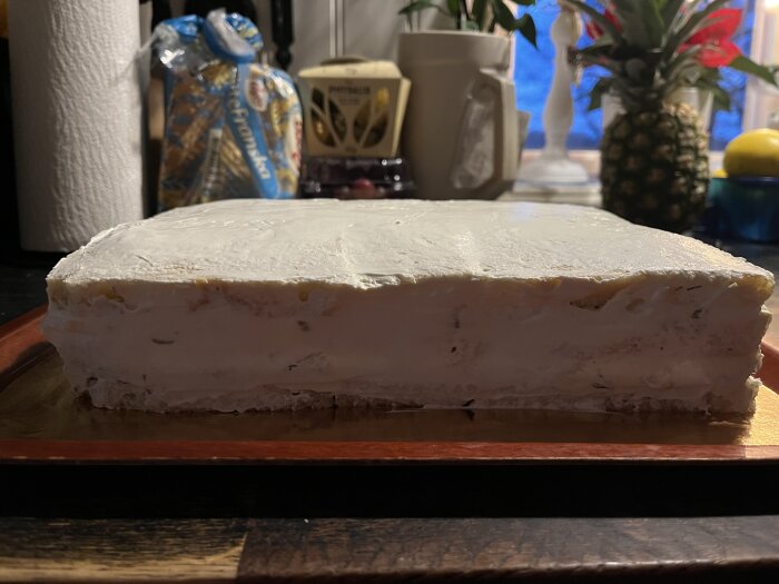 Smörgåstårta med ett lager av krämig fyllning, täckt med färskost och gräddfil, står på en mörk bricka i ett kök.