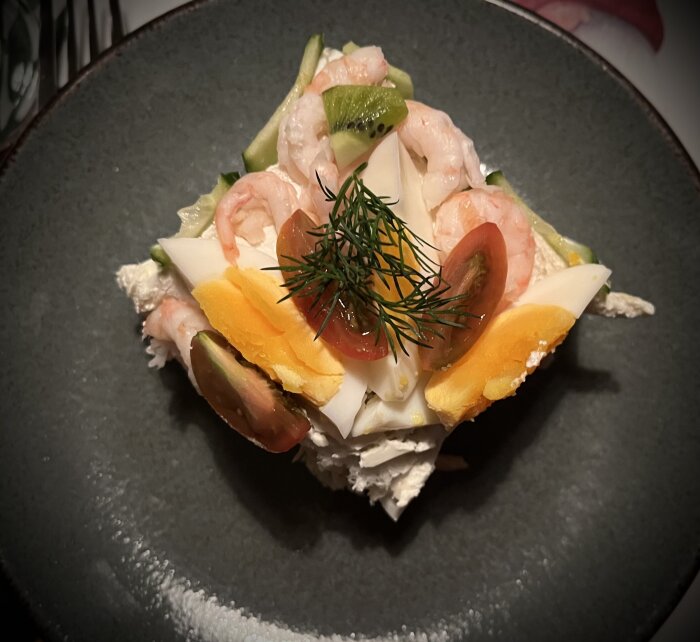 En bit smörgåstårta med räkor, ägg, dill, tomathalvor, kiwi och gurka på en mörk tallrik.