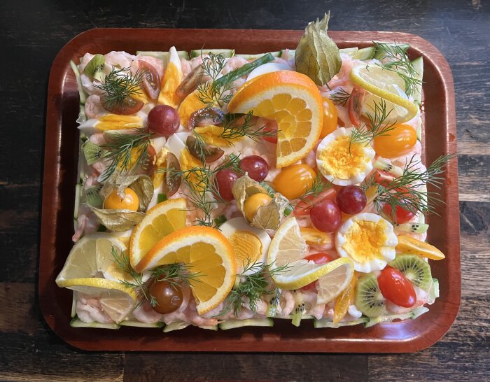 Smörgåstårta garnerad med räkor, ägg, physalis, dill, kiwi, tomater, apelsin och citron på en träbricka.