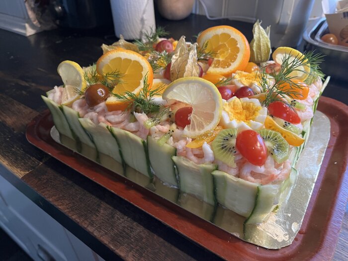 Smörgåstårta med dekorationer av skalade räkor, gurk- och citronskivor, tomater, kiwi, physalis, apelsin och dill på en bricka.