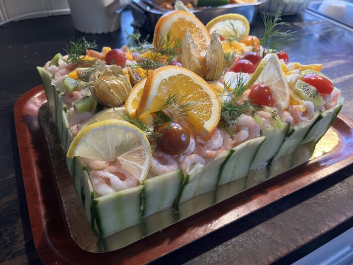 Smörgåstårta dekorerad med räkor, skivor av apelsin och citron, tomater, physalis och dill, omgiven av gurkskivor, serverad på ett fat.