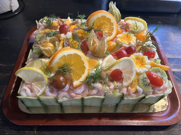 Smörgåstårta dekorerad med räkor, apelsin- och citronskivor, körsbärstomater, physalis, dill och gurkskivor på kanten, serverad på en bricka.