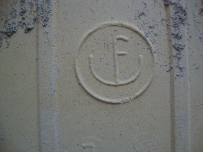 Beige kakelplatta med stämpeln F inuti en cirkel. Visar slitage och damm på ytan.