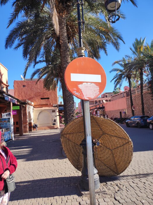 En röd trafikskylt med vit linje, förbjuden infart, står på en gata i Marrakech omgiven av palmer och terrakottabyggnader.