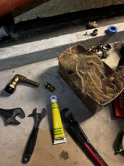 Verktyg och material för rörbyte, inklusive skiftnyckel, rörtång, gängtejp, packpasta och mässingskopplingar, ligger på ett betonggolv.