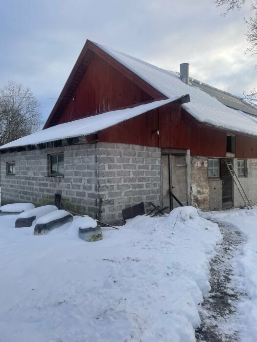 Bild på en ladugård med snötäckt tak och en vägg av olika typer av bruk, inklusive gammal kalkputs och cementputs. En stege lutar mot väggen.