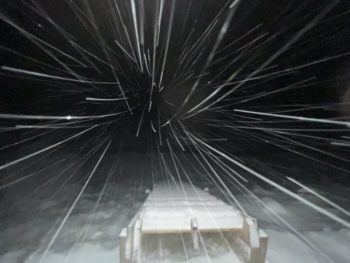 Snöstorm blixtrar över en nattlig brygga, skapar långa linjer av ljus och snöflingor i luften.