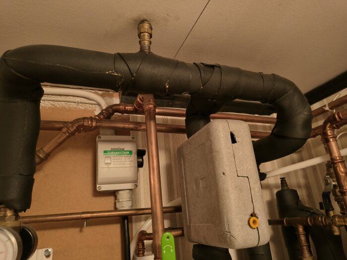 Isolerade rör och en tryckmätare i ett värmesystem, med en vit styrenhet fäst på väggen.