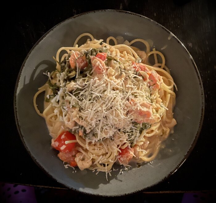 Pasta med krämig räksås, toppad med riven parmesan, räkor, tomater och grönsaker i en ljus sås på en grå tallrik.