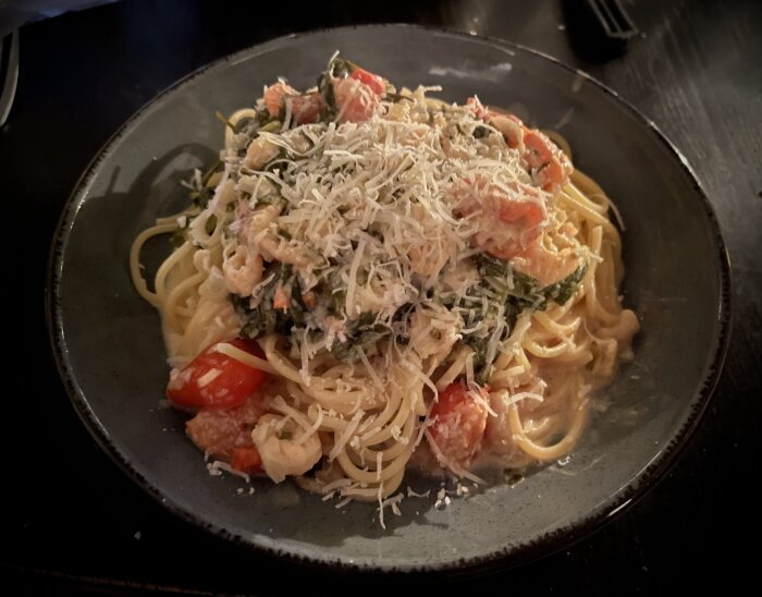 Pasta med räksås, toppad med riven parmesan, tomater och bladgrönt i en mörkgrå skål.