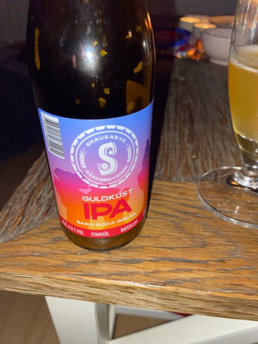 En flaska Snausarve Gårdsbryggeri Guldkust IPA med ett glas öl på ett träbord.