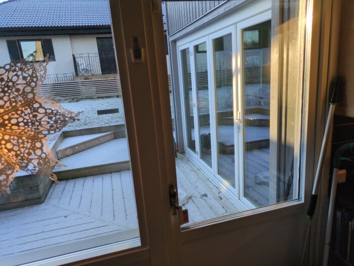 Vikparti med glasdörrar och vinklad altan utomhus belagd med tunn snö, samt ett angränsande hus med vit fasad i bakgrunden.