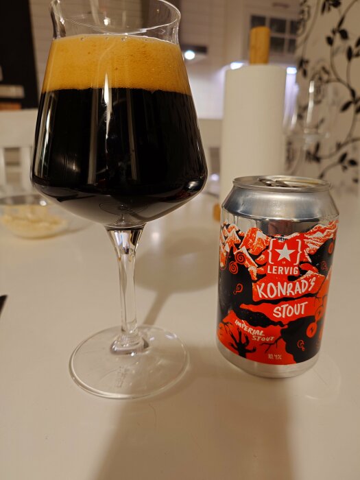 Glas med mörkt öl och skum bredvid en burk Lervig Konrad's Stout på ett vitt bord.