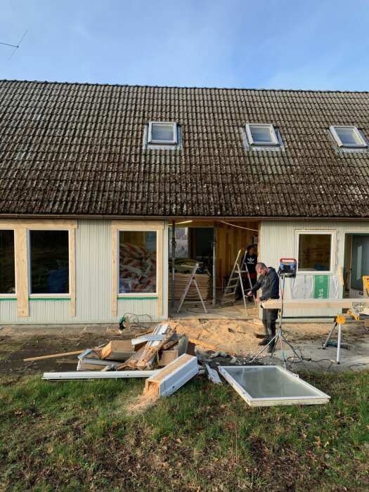 Renovering av Mockfjärd elementhus från 1969 med byggmaterial spritt på gräset och arbetare som använder verktyg framför huset.