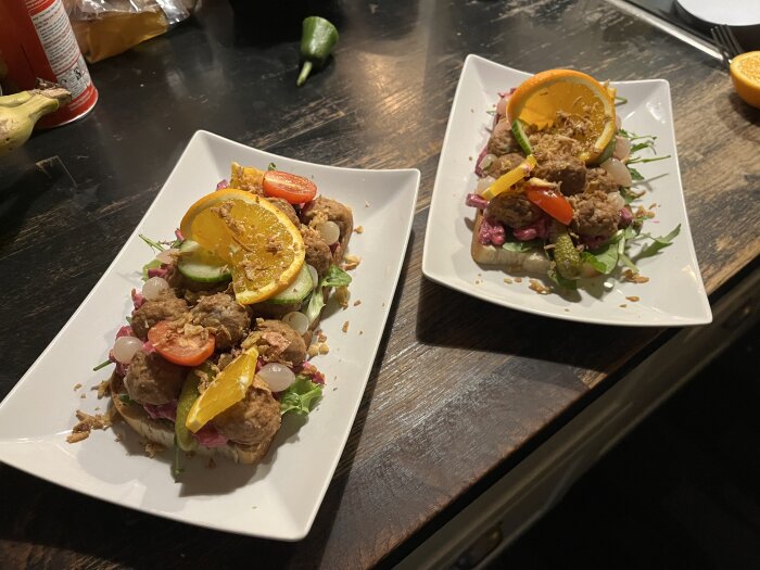 Två tallrikar med köttbullsmackor, dekorerade med orange- och tomatklyftor, gurkskivor, syrad lök och sallad på mörkt träbord.