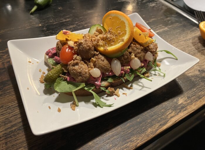 Smörgås med köttbullar, rödbetssallad, syltlök, saltgurka, tomat, apelsinskiva och krispig lök på en tallrik.