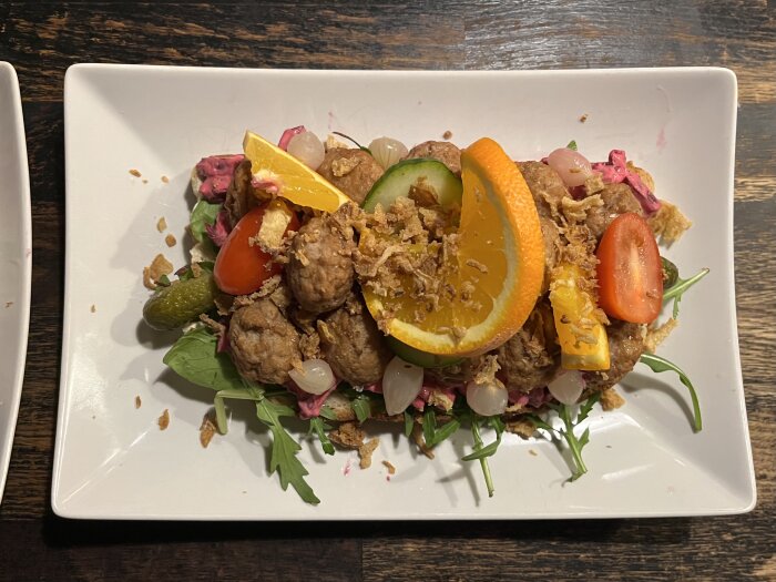 Köttbullsmacka serverad på en tallrik, toppad med rödbetssallad, tomat, gurka, apelsin, syltlök, pickles och rostad lök.