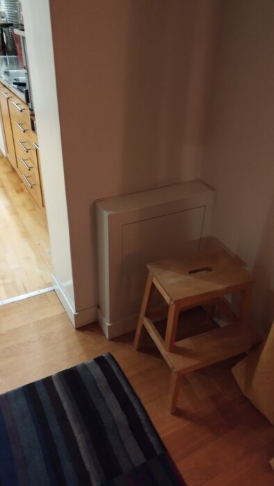 Platsbyggd vit mdf-låda som döljer en fördelare på övervåning. Placerad intill en vit vägg och bredvid en träpall på trägolv.