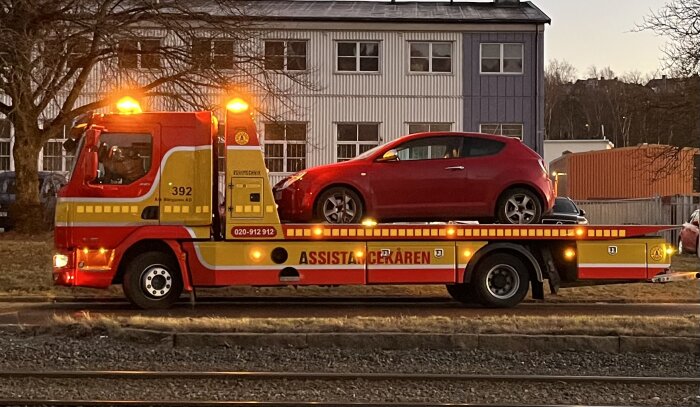 Bärgningsfordon med blinkande ljus transporterar en röd bil på flaket, parkerat vid en byggnad i skymning.