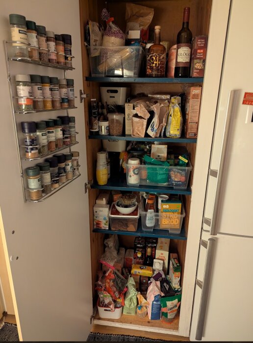 Platsbyggt skafferi med öppna dörrar, fullt med matvaror på hyllor och kryddor på dörren, plastbackar används för organisering.
