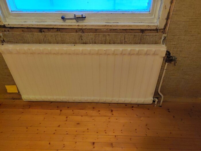 Vitt vattenburet element under fönster, anslutet med rör på höger sida, står på trägolv i rustik matsal under renovering.